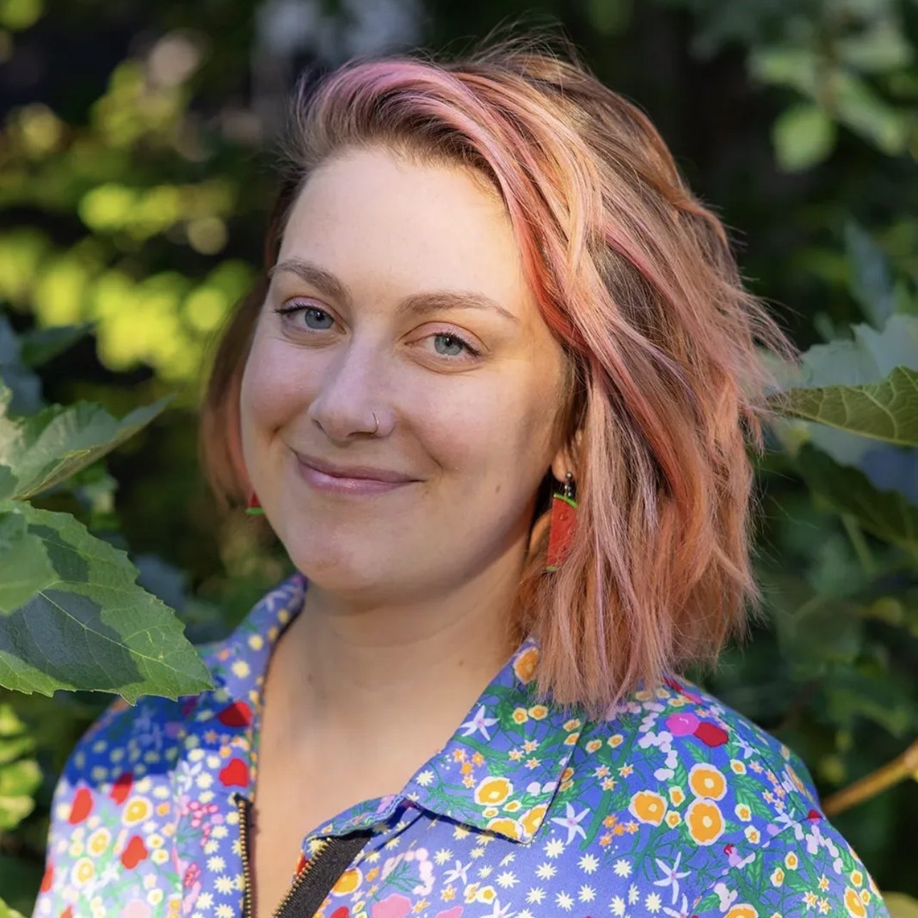 Chloe Jacobson, LPC/ATR (she/her) White woman with pinkish-red shoulder-length hair and a blue shirt with multicolored flowers stands in front of a leafy outdoor background
